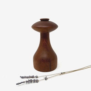 以下が含まれることがあります： キノコのようなデザインの手作りの木製ペッパーグラインダー。 グラインダーは濃い茶色の木材でできており、丸みを帯びたベース、細い首、平らな円盤状のトップが付いています。 2本のラベンダーの小枝がグラインダーの近くに置かれています。
