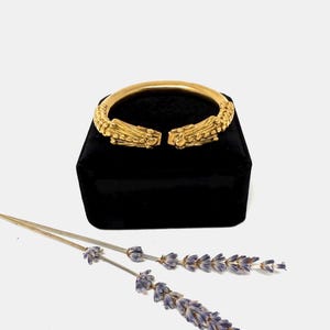 May include: A gold-colored cuff bracelet with ornate, textured detailing. The bracelet is displayed on a black velvet box, with dried lavender sprigs in the foreground. The bracelet's design features intricate patterns and a clasp closure.