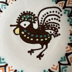May include: A white ceramic plate with a brown rooster design. The rooster has a green and yellow crest and tail feathers. The plate is decorated with a brown, orange, and green geometric border.