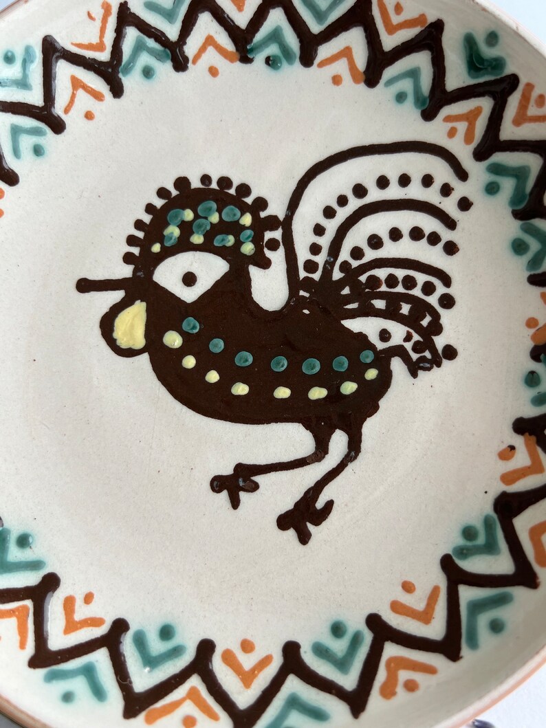 May include: A white ceramic plate with a brown rooster design. The rooster has a green and yellow crest and tail feathers. The plate is decorated with a brown, orange, and green geometric border.