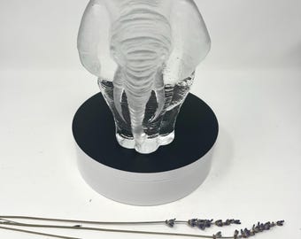 May include: A clear glass sculpture of an elephant standing on a black and white pedestal. The elephant is facing forward with its trunk down.
