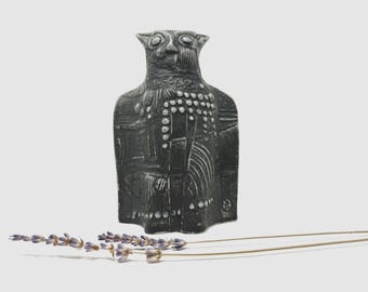 Könnte beinhalten: Eine dunkelgraue, stilisierte Eulen-Skulptur mit strukturierter Oberfläche. Die Eule hat große Augen und ein detailliertes Federmuster. Kleine weiße Punkte schmücken den Brustbereich. Ein Zweig Lavendel liegt im Vordergrund.