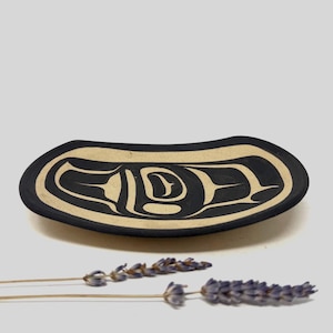 May include: A black and beige decorative dish with a Native American design. The dish has an oval shape with a black exterior and a beige interior featuring a stylized design. Dried lavender sprigs are in the foreground.