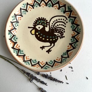May include: A white ceramic plate with a brown rooster design. The rooster has a green and yellow crest and tail feathers. The plate has a brown, orange, and green geometric border.