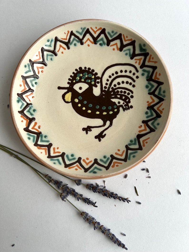 May include: A white ceramic plate with a brown rooster design. The rooster has a green and yellow crest and tail feathers. The plate has a brown, orange, and green geometric border.