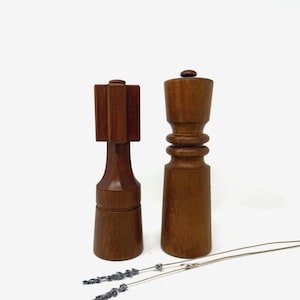 以下が含まれることがあります： 木製の塩とコショウ挽き器が2つ。左側の挽き器は長方形の上面を持ち、右側の挽き器は丸みを帯びた上面を持っています。どちらも濃い色の木材でできており、白い表面に置かれています。乾燥したラベンダーの小枝が前景にあります。