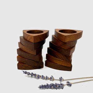 May include: Two wooden napkin rings with a triangular top and stacked, curved sides. The napkin rings are a rich brown colour and are next to sprigs of lavender.