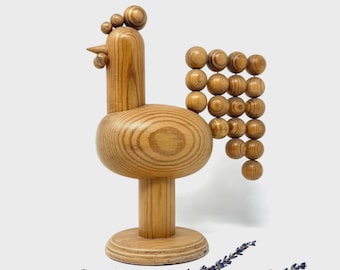 May include: A wooden rooster sculpture with a rounded body, a long neck, and a circular base. The rooster's tail is made of wooden spheres. The sculpture is light brown and sits on a white surface. Lavender sprigs are in the foreground.