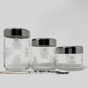 Könnte beinhalten: Drei transparente Glasbehälter mit silbernen Deckeln, jeweils in unterschiedlicher Größe. Die Gläser sind mit einem sich wiederholenden Muster aus weißen Strichmännchen verziert. Ein Zweig getrockneten Lavendels liegt vor den Gläsern.