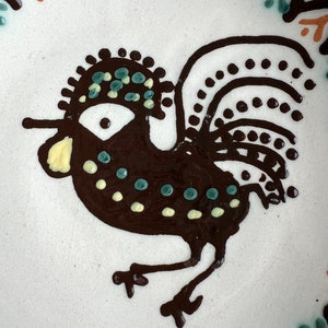 May include: A brown rooster with a yellow beak and black and white polka dots on its body. The rooster has a green and blue crest and a black tail with white polka dots.