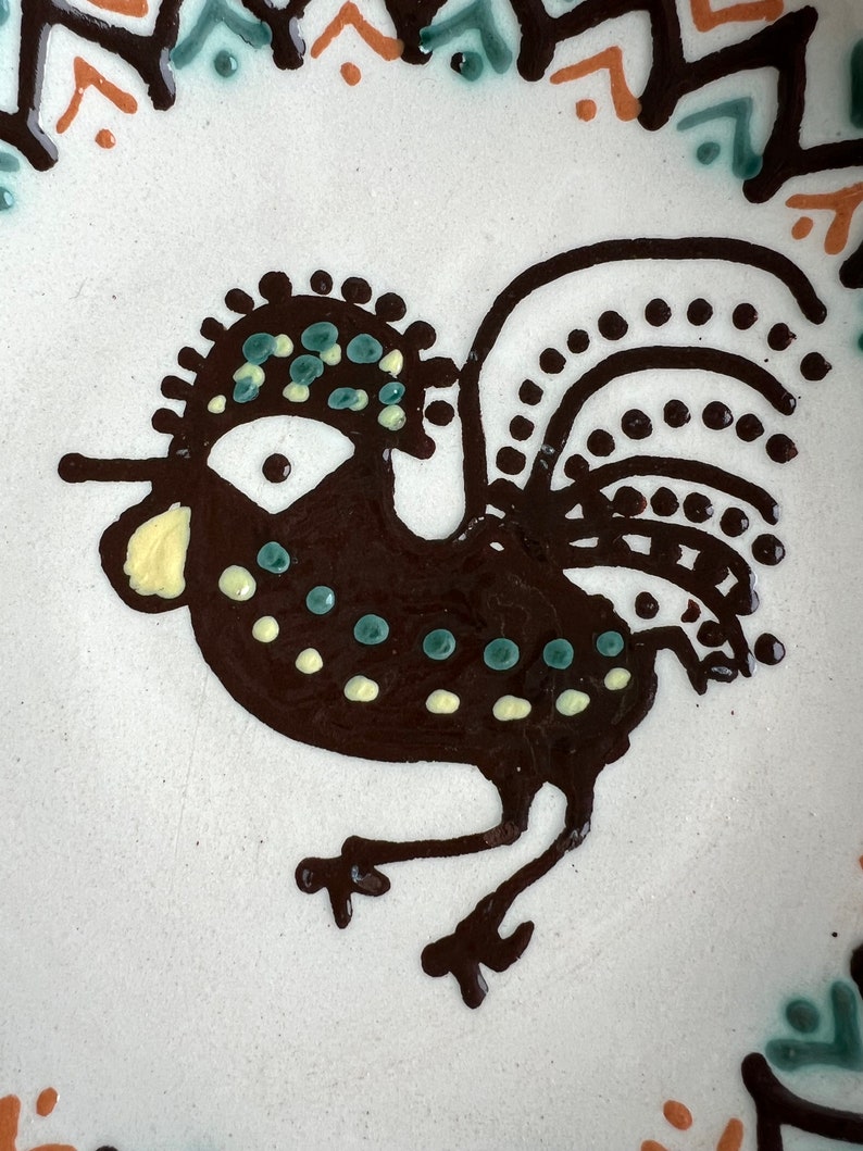 May include: A brown rooster with a yellow beak and black and white polka dots on its body. The rooster has a green and blue crest and a black tail with white polka dots.