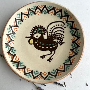 May include: A white ceramic plate with a brown rooster design. The rooster has a yellow beak, green eyes, and brown and black feathers. The plate has a brown, orange, and green geometric border.