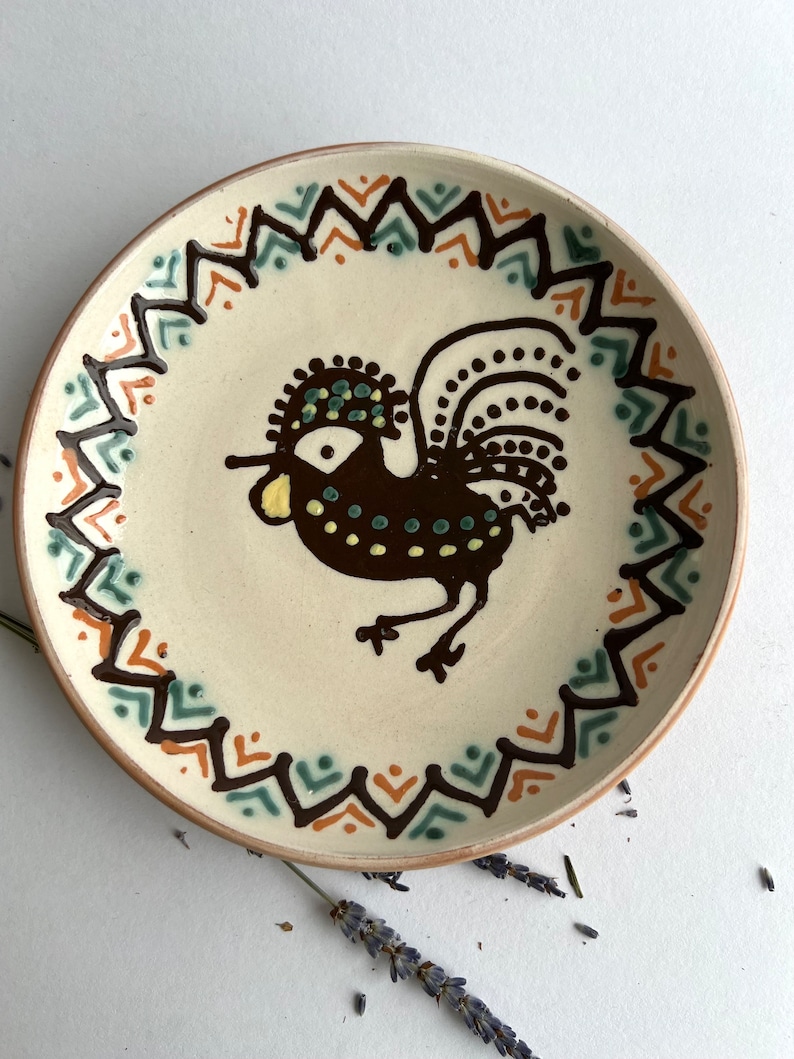 May include: A white ceramic plate with a brown rooster design. The rooster has a yellow beak, green eyes, and brown and black feathers. The plate has a brown, orange, and green geometric border.