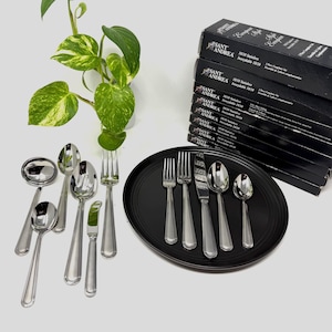 May include: A collection of stainless steel silverware, including forks, knives, and spoons, arranged on a black oval plate. Several boxes of silverware are stacked in the background. A green plant with patterned leaves is in the background.
