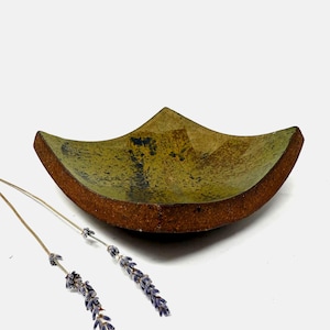 May include: A handmade ceramic bowl with a unique, angular design. The bowl features a green interior with dark accents and a textured, brown rim. Two sprigs of lavender are placed near the bowl.
