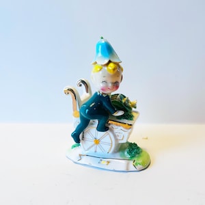 Puede incluir: Una figurita de porcelana vintage de un niño sonriente que lleva un sombrero azul, empujando un carro blanco con flores verdes y detalles dorados. La figurita está sobre una base blanca con hierba verde.