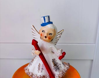 May include: A white porcelain angel figurine with blue and white striped hat, holding a red wand. The angel is standing on a small orange plate with white polka dots and the words "JULY ANGEL" written on it.
