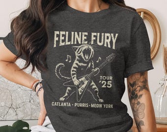 Puede incluir: Camiseta gris oscuro con un diseño gráfico vintage de un gato tocando la guitarra, con el texto "Feline Fury Tour '25" y nombres de ciudades. El diseño es de estilo vintage.