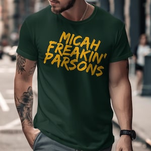 Puede incluir: Camiseta verde oscuro con el texto amarillo "MICAH FREAKIN' PARSONS" en diagonal. La camiseta es de cuello redondo y parece estar hecha de un material suave. El diseño es un gráfico audaz y llamativo.