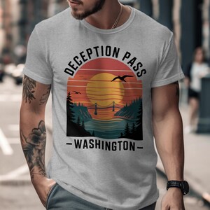 May include: A light gray t-shirt featuring a sunset design with a bridge over water, trees, and flying birds. The text "DECEPTION PASS - WASHINGTON" is printed in black. The shirt is short-sleeved.