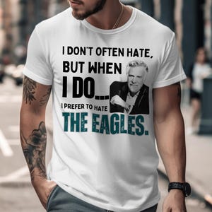 Könnte beinhalten: Weißes T-Shirt mit dem Text "I DON'T OFTEN HATE, BUT WHEN I DO... I PREFER TO HATE THE EAGLES." Das Shirt zeigt ein Schwarz-Weiß-Bild eines Mannes. Der Text "THE EAGLES" ist türkis.