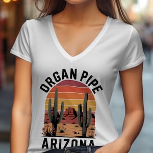 May include: White V-neck t-shirt featuring a desert landscape design with the text "ORGAN PIPE ARIZONA". The graphic includes cacti, mountains, and a sunset color scheme.