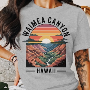 May include: A light gray t-shirt featuring a colorful graphic of Waimea Canyon in Hawaii. The design includes a sunset, canyon landscape, and the words "WAIMEA CANYON" and "HAWAII".