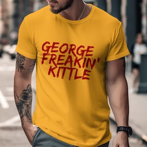 Puede incluir: Camiseta de color amarillo mostaza con el texto "GEORGE FREAKIN' KITTLE" en rojo, con letras estilo pincel. La camiseta tiene mangas cortas y cuello redondo. La imagen está tomada al aire libre.
