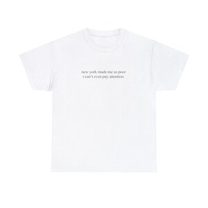 Op de afbeelding: Wit T-shirt met de tekst "new york made me so poor i can't even pay attention."