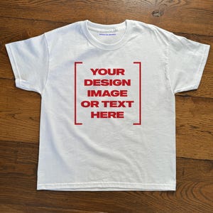 Puede incluir: Una camiseta blanca con un marco rectangular rojo en la parte delantera. El marco contiene el texto "YOUR DESIGN IMAGE OR TEXT HERE" en letras rojas. La camiseta se muestra sobre una superficie de madera.