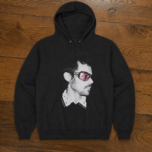 May include: Black zip-up hoodie with a portrait of a person wearing pink sunglasses. The black and white portrait is on the front of the hoodie. The hoodie has a drawstring hood and a front pocket. The person is wearing a collared shirt.
