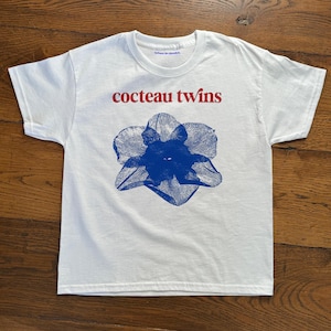 May include: White t-shirt featuring the text "cocteau twins" in red above a blue floral design. The shirt has a crew neck and short sleeves. The text "when in doubt." is printed on the collar.