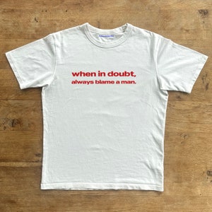 May include: White t-shirt with red text that reads "when in doubt, always blame a man."