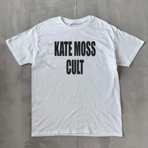 May include: White t-shirt featuring the words "KATE MOSS CULT" in bold black lettering. The top has a crew neck and short sleeves. The garment is displayed flat on a grey surface.