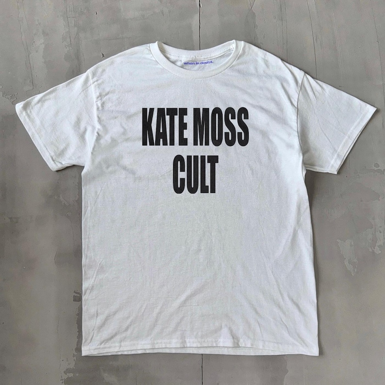 May include: White t-shirt featuring the words "KATE MOSS CULT" in bold black lettering. The top has a crew neck and short sleeves. The garment is displayed flat on a grey surface.