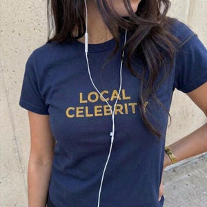 May include: A navy blue short-sleeved t-shirt featuring the text LOCAL CELEBRITY printed in a distressed gold font across the chest. A thin white wired earphone cord hangs down the center of the shirt from the neckline.