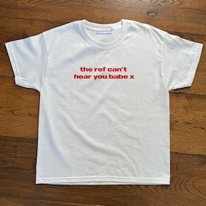 以下が含まれることがあります： 赤い文字で「the ref can't hear you babe x」と書かれた白いTシャツ。柔らかい素材でできており、クルーネックです。テキストはシャツの前面の中央に配置されています。
