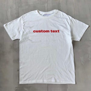 Op de afbeelding: Wit T-shirt met de tekst "custom text" in rood gedrukt op de voorkant.