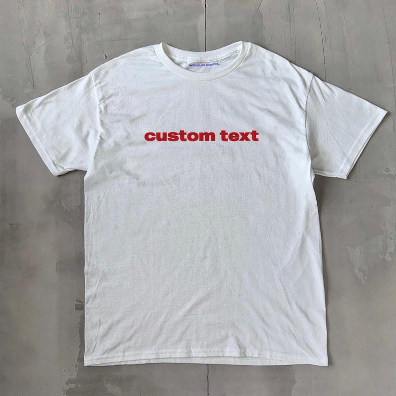 Op de afbeelding: Wit T-shirt met de tekst "custom text" in rood gedrukt op de voorkant.