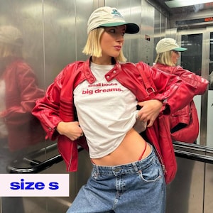 May include: A red leather jacket with a white t-shirt that reads "small boobs, big dreams." The model is wearing blue jeans and a beige baseball cap with a green brim. The image also shows the text "size s".