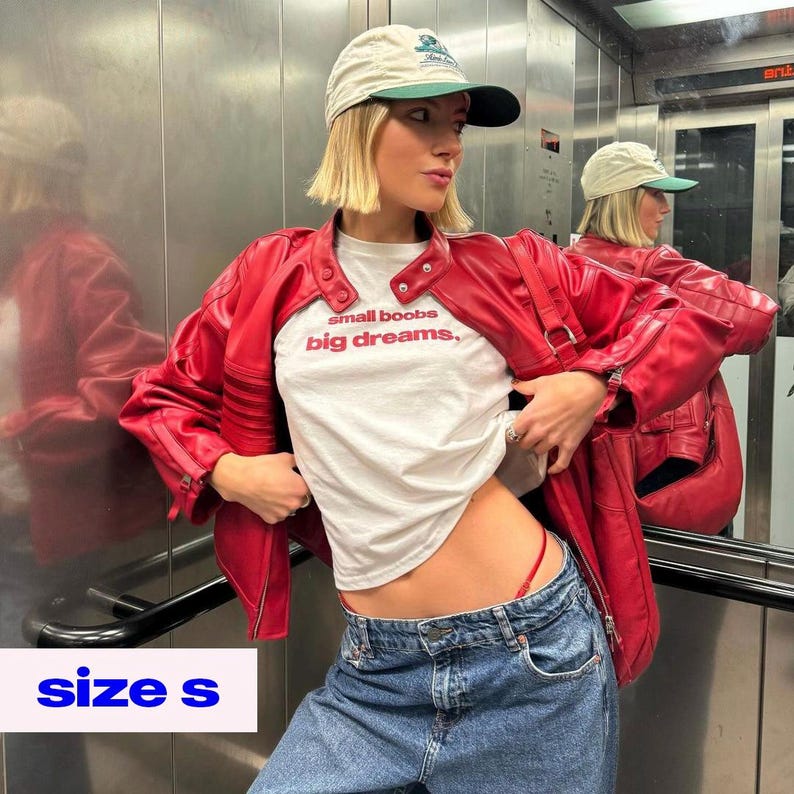 May include: A red leather jacket with a white t-shirt that reads "small boobs, big dreams." The model is wearing blue jeans and a beige baseball cap with a green brim. The image also shows the text "size s".