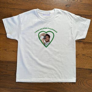 Può includere: T-shirt bianco con la scritta verde "I love my Irish boyfriend". Un cuore verde contiene una fotografia di un uomo che tiene una macchina fotografica. La maglietta ha girocollo e maniche corte.