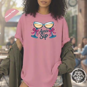 May include: A pink t-shirt with a graphic of two champagne glasses with blue stems and pink bows. The text "Après Sip" is printed below the glasses in teal. The shirt is made of 100% cotton.