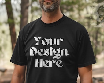 Op de afbeelding: Een zwart T-shirt met de tekst "Your Design Here" in wit gedrukt op de voorkant. Het shirt wordt gedragen door een persoon die in een bos staat.