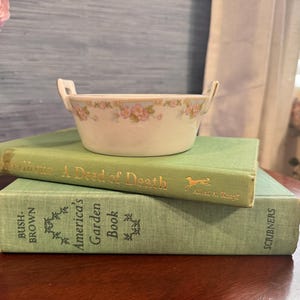 May include: A small, off-white ceramic bowl with two handles, adorned with a floral pattern in pink and green. The bowl rests on two vintage green books. The top book reads "A Deed of Death."