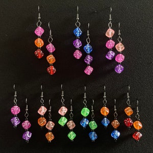 May include: A collection of colorful dice earrings. Each earring features three translucent dice in a gradient of colors, including pink, orange, green, and blue. The dice are strung together with black beads and silver-tone hooks.