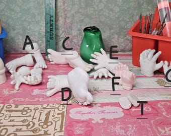 Può includere: Una collezione di calchi in gesso bianco di mani, piedi e altre parti del corpo, insieme a strumenti per scolpire e un vaso di vetro verde. I calchi sono disposti su una superficie rosa a fantasia. È visibile una cassetta degli attrezzi blu.