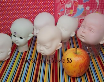 May include: A collection of doll heads in various stages of completion, including a pale green elf-like head and several blank baby doll heads. An apple rests in the foreground. The background features a striped fabric and a pink patterned surface.