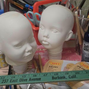May include: Two white, unpainted baby doll heads sit on a table with art supplies. A ruler with text reading "237 East Olive Avenue Burbank, Calif." is in the foreground. Other items include clay, paintbrushes, and a spray bottle.