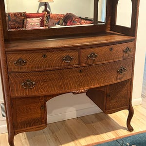 May include: Antique wooden vanity with a mirror and drawers. The vanity has a curved front with ornate hardware and a warm, reddish-brown finish. The mirror is rectangular with a small arched mirror on the side. The vanity sits on curved legs.